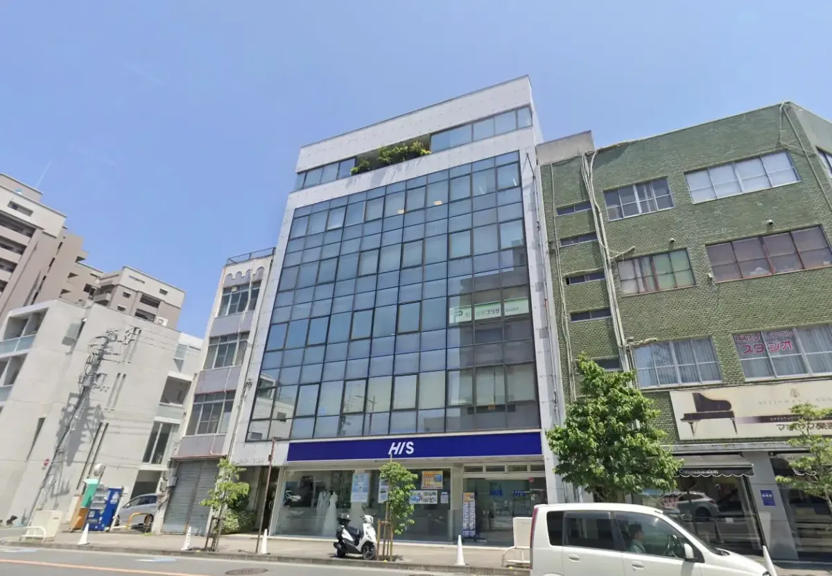 ガラス張りの中層ビルの外観。1階にHISの店舗が入っており、前の道路には白い車とバイクが停まっている。晴天の青空の下、両隣には異なるデザインのビルが並んでいる街並みの写真。